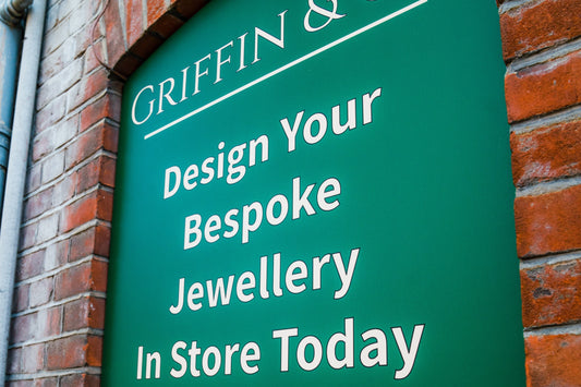 A close-up of a green sign with white text partially visible, reading "GRIFFIN & Co. Design Your Bespoke Jewellery In Store Today," mounted on a brick wall.