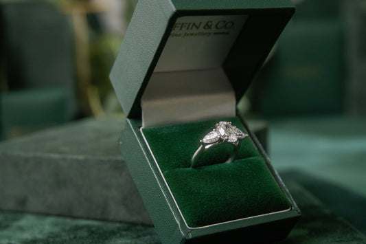 An open dark green ring box showcasing a brilliant three-stone diamond engagement ring, set against a blurred background of green velvet.