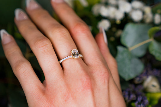 A close-up of a hand wearing a bespoke radiant-cut diamond engagement ring set in yellow gold, with a pavé diamond band, against a soft-focus background of green foliage and white flowers.