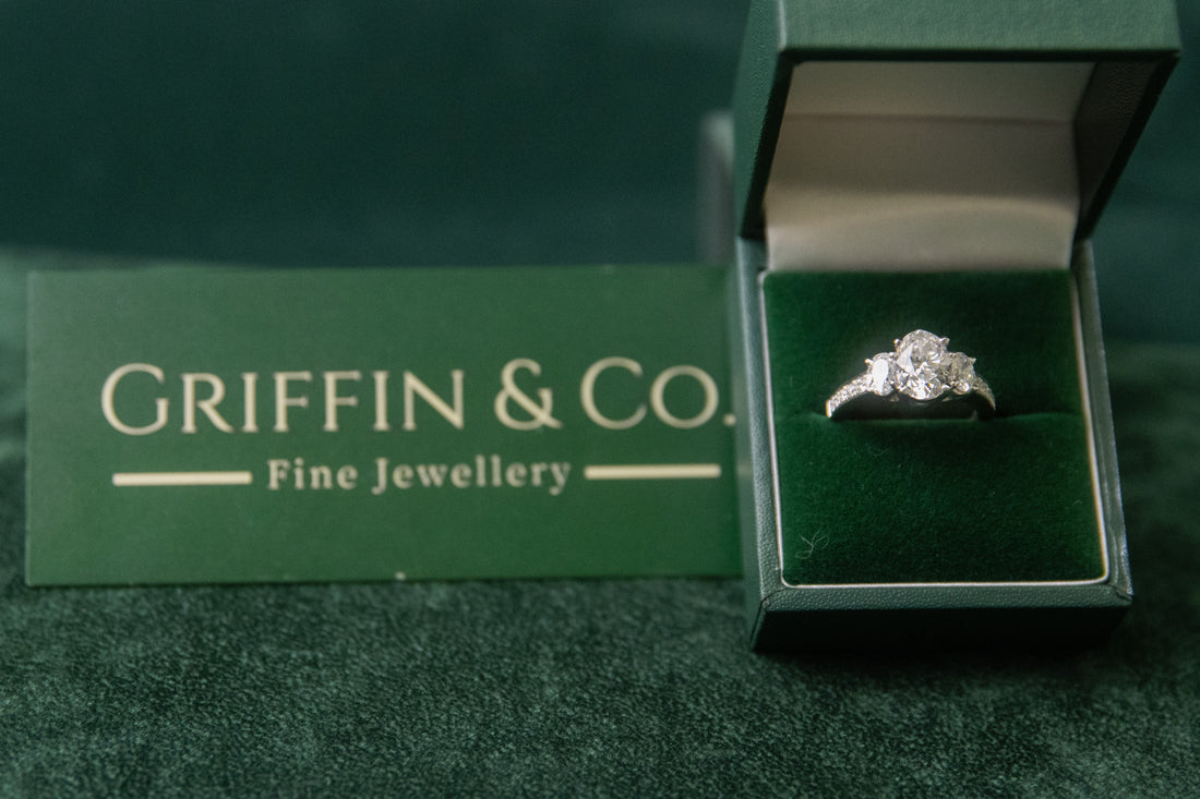 An open green ring box displaying a sparkling three-stone diamond ring with a halo setting, resting on a dark green textured surface next to a "Griffin & Co. Fine Jewellery" branded card.