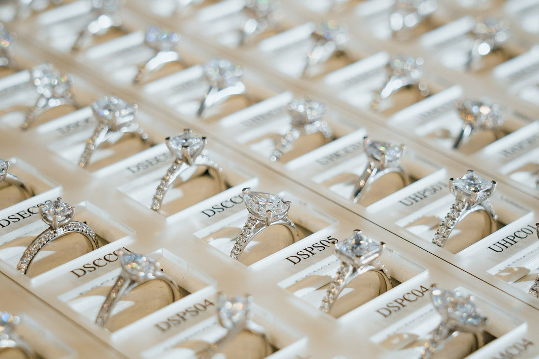 A close-up photograph of a display tray filled with numerous lab-grown diamond engagement rings. The rings are arranged in neat rows, showcasing various styles and diamond cuts.