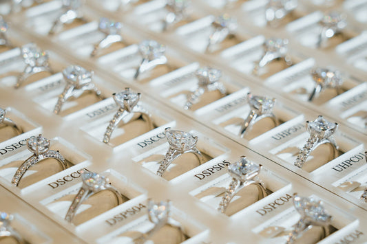 A close-up photograph of a display tray filled with numerous lab-grown diamond engagement rings. The rings are arranged in neat rows, showcasing various styles and diamond cuts.