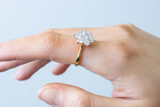 A close-up of a hand wearing a vintage-style cluster ring featuring multiple diamonds set in a flower shape on a yellow gold band.