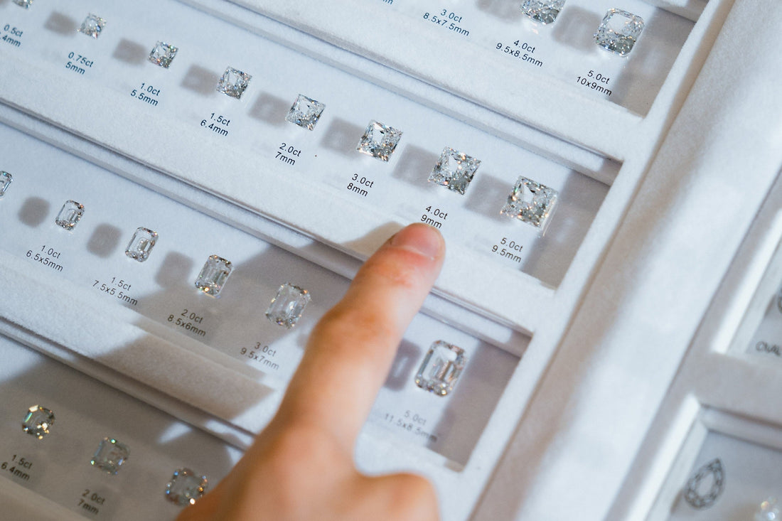 A close-up of a person's finger pointing at a 4.0 carat lab-grown diamond in a display case. The case shows various diamond sizes, each labelled with its carat weight and dimensions, illustrating the concept of carat.