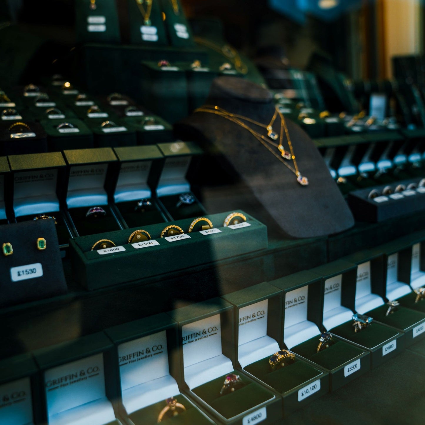 Elegant jewelry, including layered necklaces and individually boxed rings with pricing, presented in a store window.