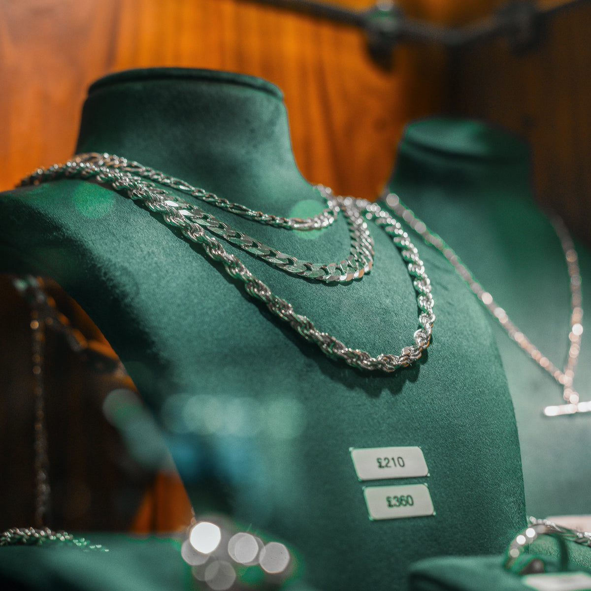 Silver chain necklaces displayed on dark green velvet jewelry busts in a retail setting, with a wooden wall background and price tags visible.
