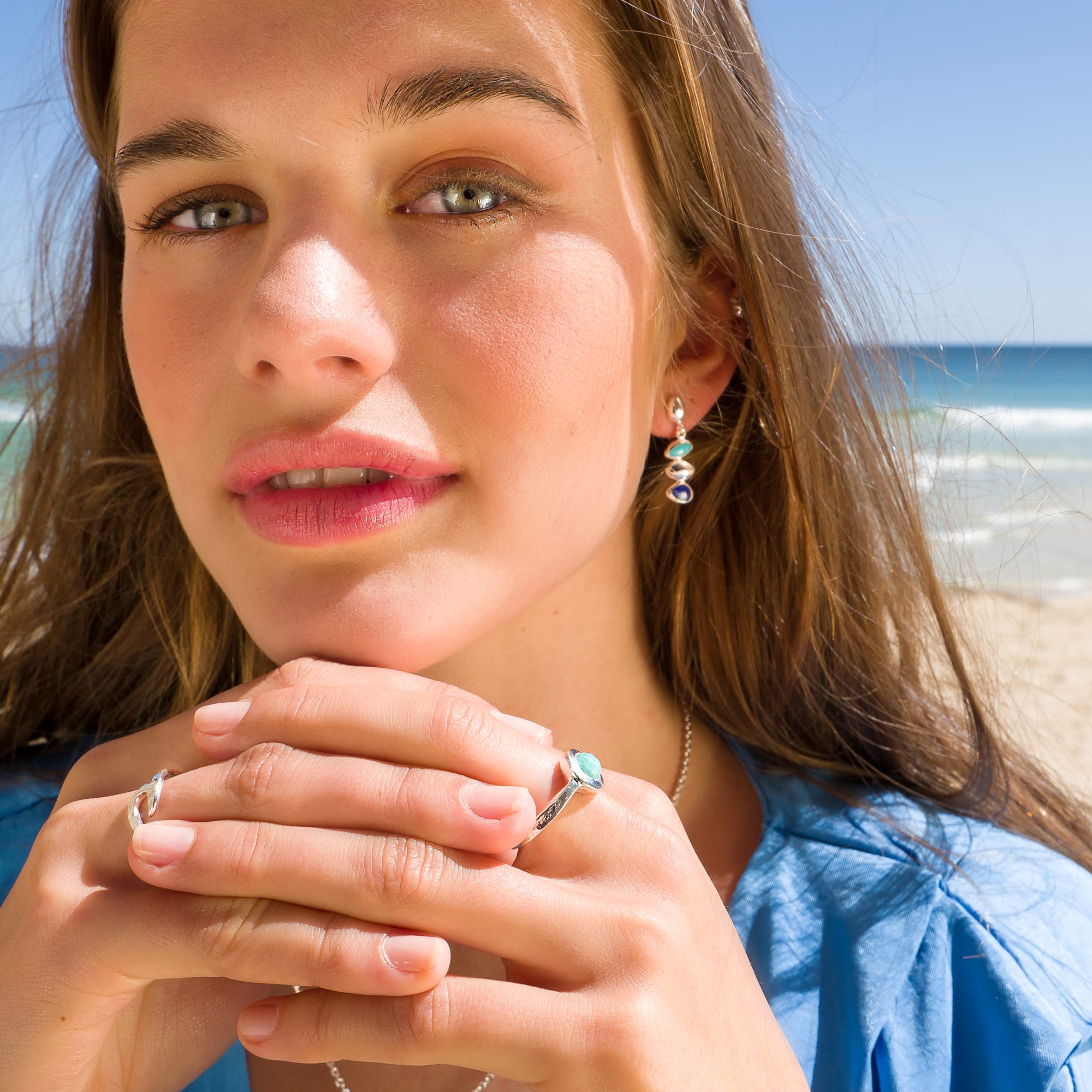 A close-up portrait of a woman on a beach wearing silver pebble stud drop earrings with turquoise and lapis stones, and matching rings.