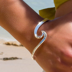 A close-up of the silver wave bangle being worn on a person's wrist at the beach, with sunlight glinting off its surface.