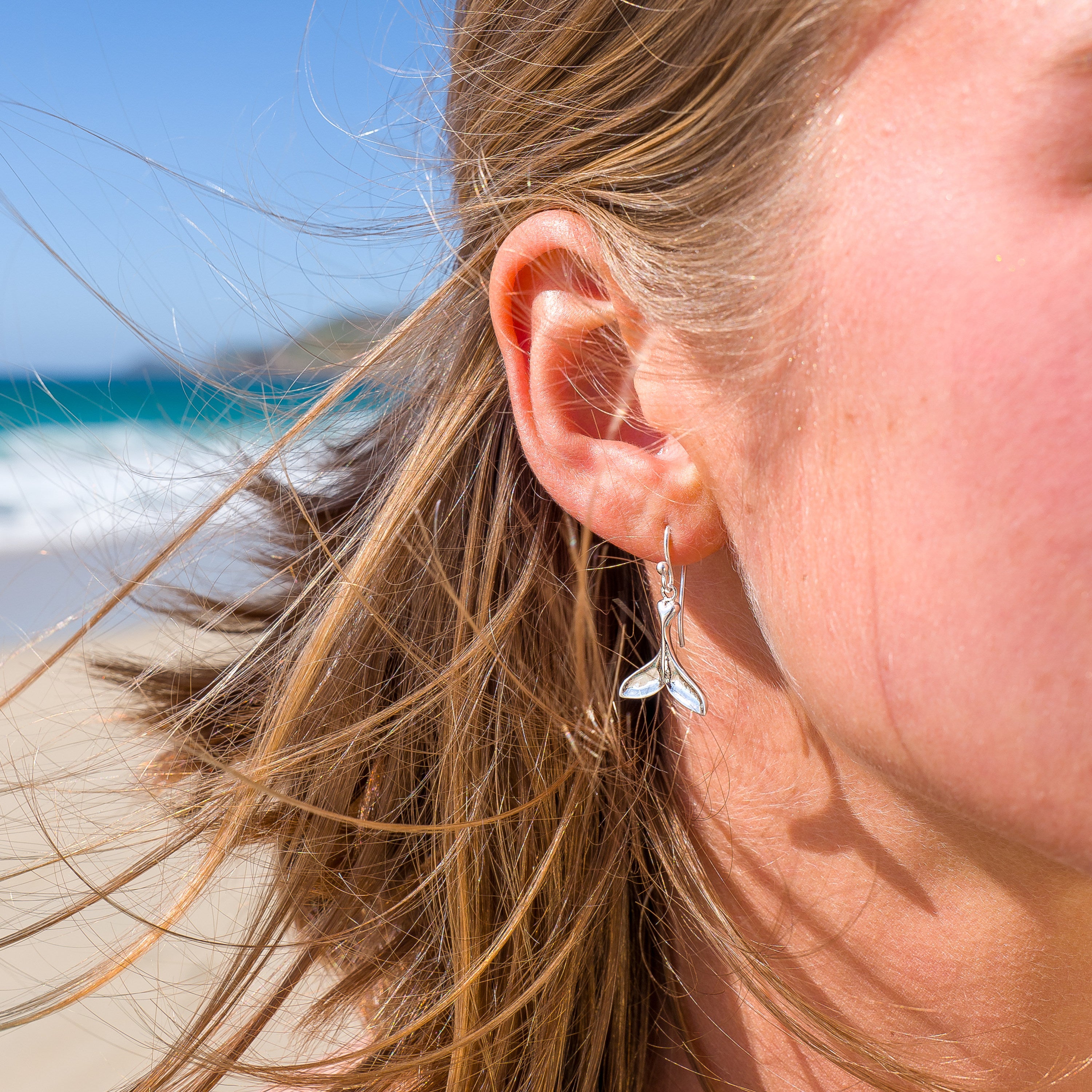 A close-up side profile of a woman on a sunny beach wearing silver whale tail drop earrings.