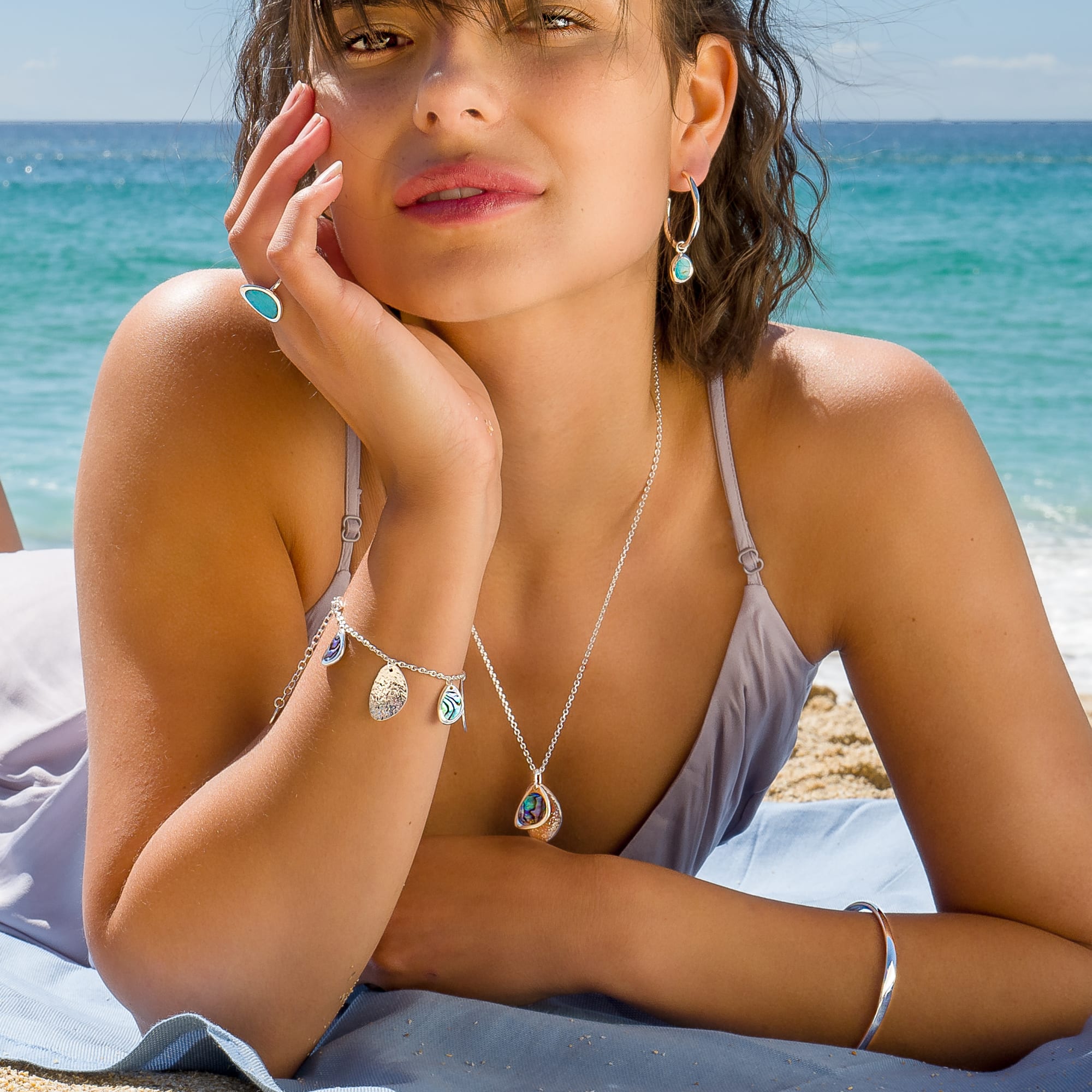  A person's arm wearing the silver bangle while lying on a sandy beach. Other jewelry, including a necklace, charm bracelet, and earrings, are also visible.