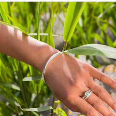 A person's arm wearing a smooth, polished silver bangle. They are holding their hand up to a green plant, with a blurred background.