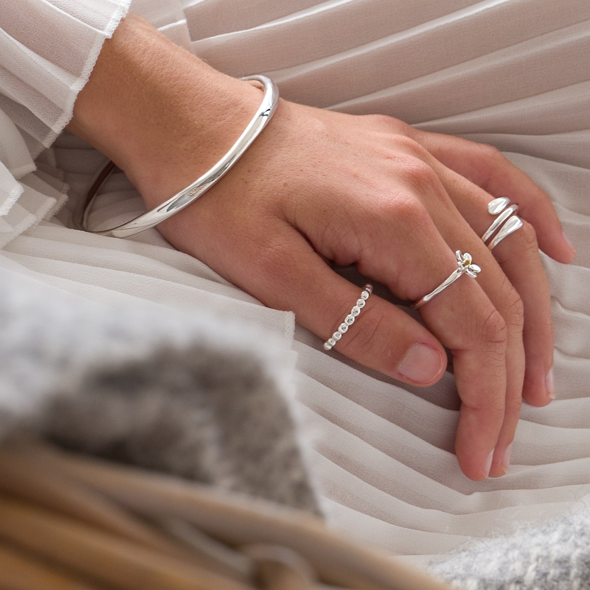 A person's hand resting on a light-colored blanket, wearing a polished silver bangle and two silver rings with simple designs.