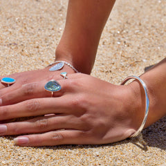 A close-up of a person's hands on a sandy beach. They are wearing a polished silver bangle and several rings on their fingers.