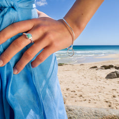 A person's arm wearing a silver bangle with a large wavy shape and two small, interlinked circles. The person is standing on a sandy beach with the ocean in the background.
