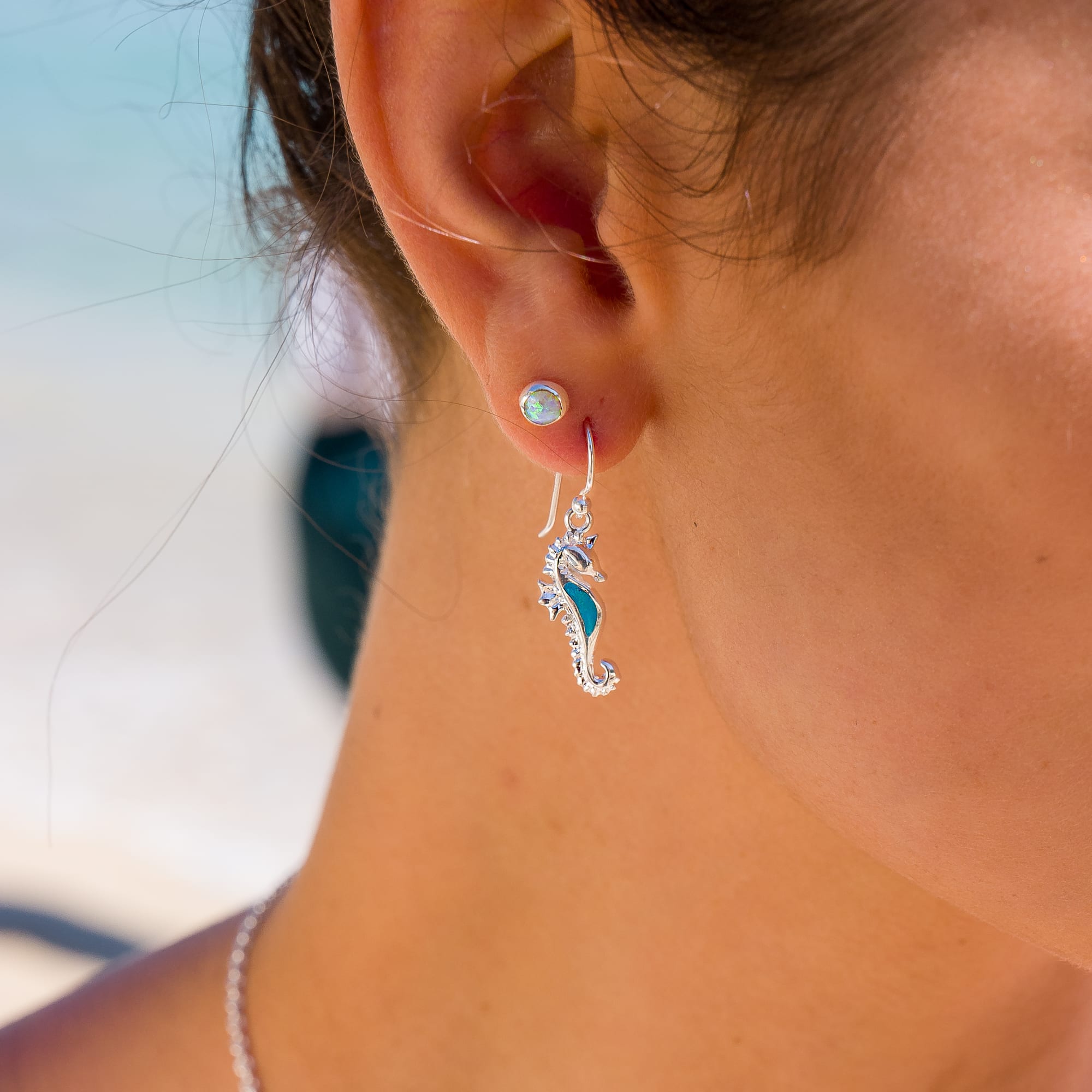 Model wearing silver seahorse hook earrings with turquoise detail against a coastal background.