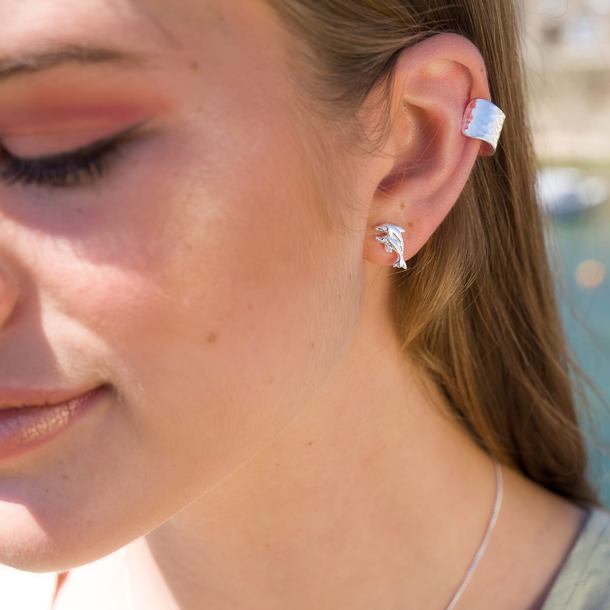 A close-up of a woman's ear wearing silver dolphin stud earrings and a silver ear cuff.