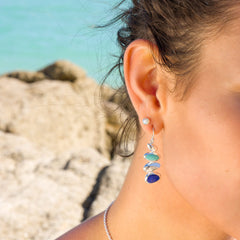 A close-up side profile of a woman wearing silver pebble drop earrings with turquoise and lapis stones, against a blurred coastal background.