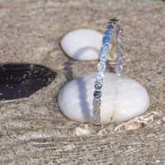 The silver pebble bangle is propped up on a smooth white stone on a sandy beach. Small ripples of water are flowing around the bangle and stone.