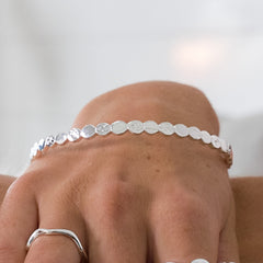 A close-up shot of a person's wrist wearing the silver pebble bangle. You can clearly see the alternating smooth and textured discs. A couple of silver rings are also visible on their fingers.