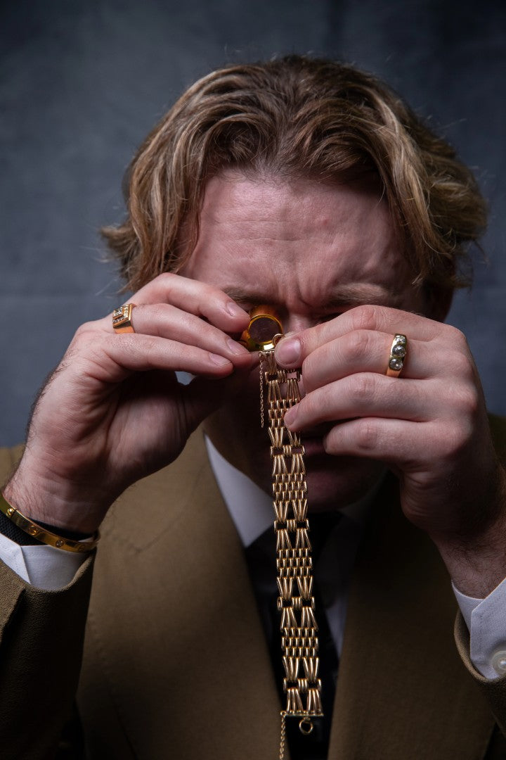 A close-up lifestyle shot of a man wearing a brown suit and white shirt, holding a gold chain necklace up to his eye with both hands, which are also adorned with gold rings.