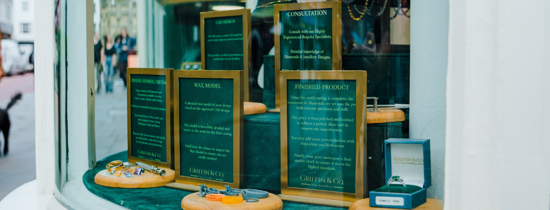 A wide shot of the Griffin & Co. jewellery shop window. The display features framed informational plaques describing the bespoke jewellery process, including "CONSULTATION," "WAX MODEL," and "FINISHED PRODUCT." Various tools and wax models are arranged on wooden platforms, with a finished ring box visible on the right. Pedestrians and a dog are blurred in the background on the street.