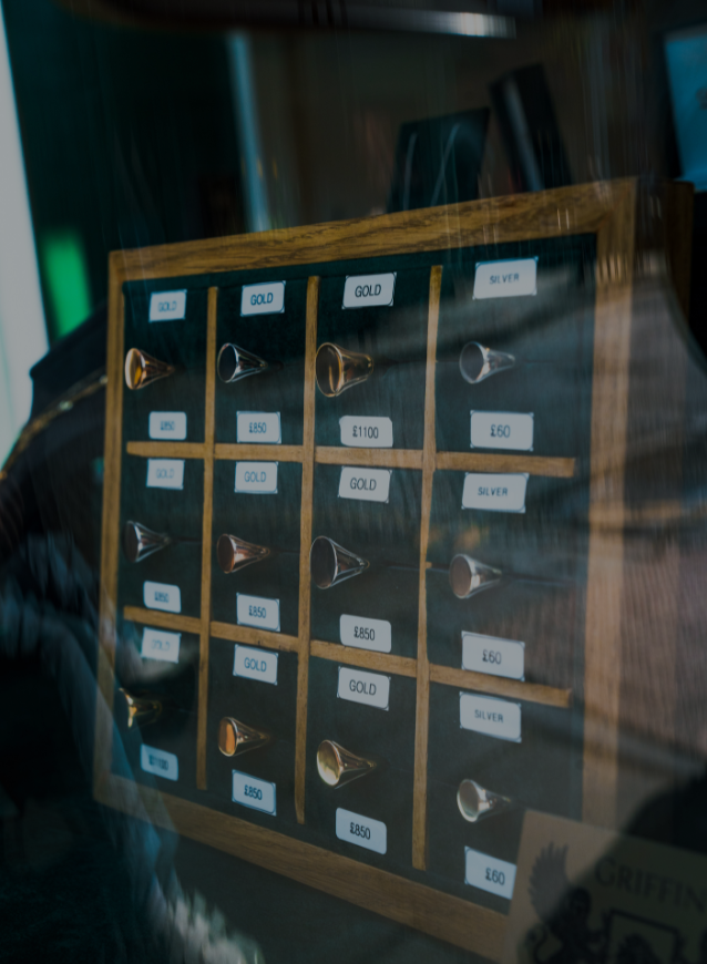 A close-up, cropped view of a wooden display stand inside a jewellery shop window. The stand holds a dark, velvet-like tray with multiple signet ring designs in different metals like gold and silver. Each ring has a small tag with a price and material. The reflections of the street are visible in the glass.