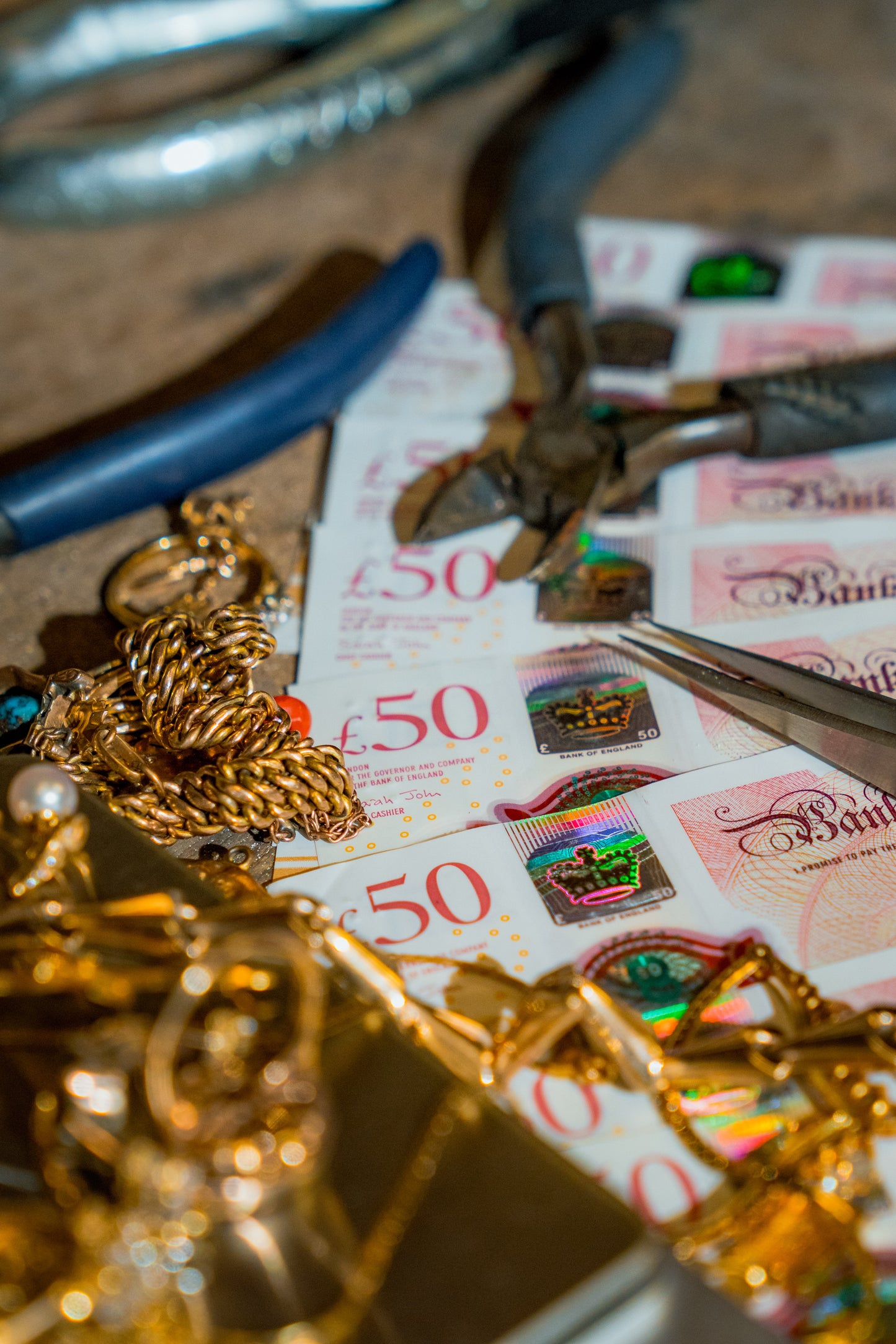 A close-up, shallow depth-of-field shot of a jewelry recycling or alteration workspace. Gold-toned jewelry, including a chunky chain and rings, is mixed with British £50 banknotes and metal pliers on a workbench.