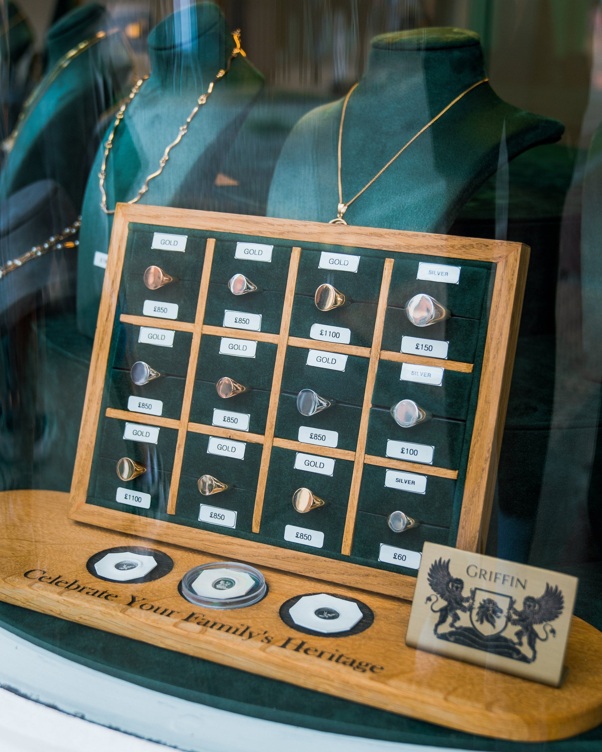 A close-up shot of a wooden display tray in a jewellery shop window. The tray holds a grid of various signet rings, showcasing different styles and metals, each with a price tag. The display rests on a wooden base with the engraved text "Celebrate Your Family's Heritage." In the foreground, a tile with the Griffin and Co. logo is visible.