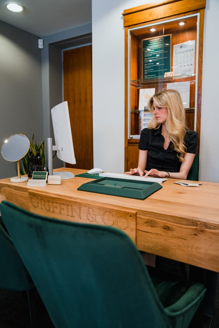 A woman with blonde hair sits behind a wooden reception desk in an office setting, with a teal velvet chair in the foreground and a wood-paneled wall in the background.