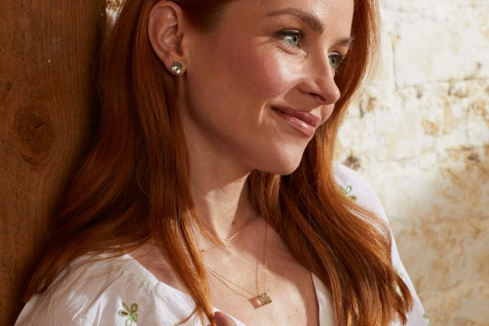 Woman with red hair wearing earrings and a necklace, standing against a wooden wall.