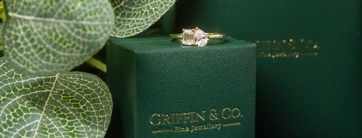 A bespoke 'Toi et Moi' lab-grown diamond engagement ring in yellow gold, featuring two unique diamond cuts. This ethical fine jewellery piece by Griffin & Co, Chichester, is elegantly displayed on its signature green ring box.