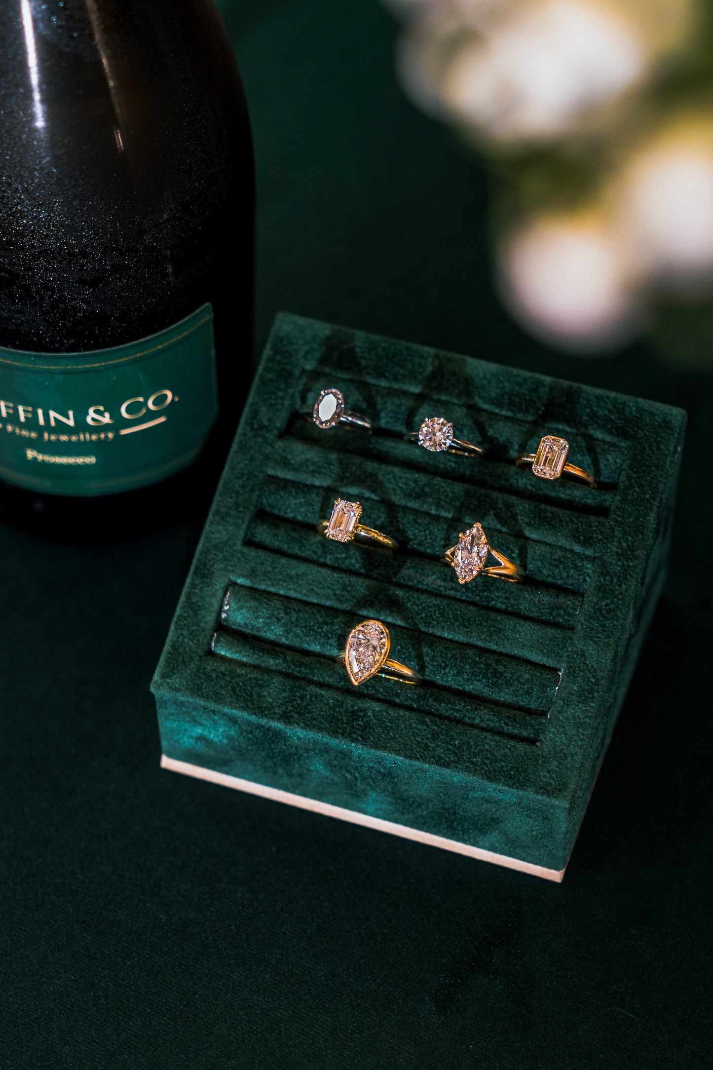 Green velvet jewellery display with gold engagement rings next to a bottle of champagne on a dark surface.