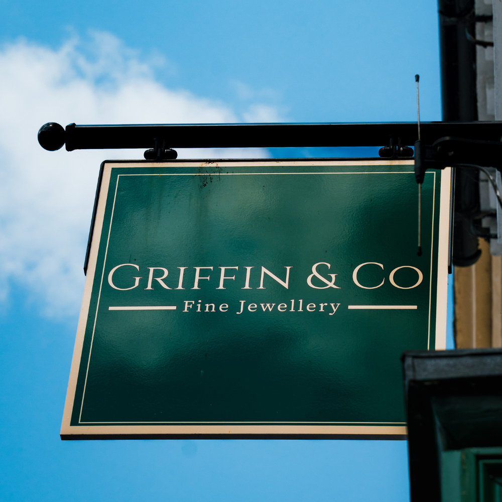 The dark green hanging sign for Griffin & Co Fine Jewellery, viewed from a low angle against a blue sky with faint clouds. The sign is suspended from a black metal bracket.