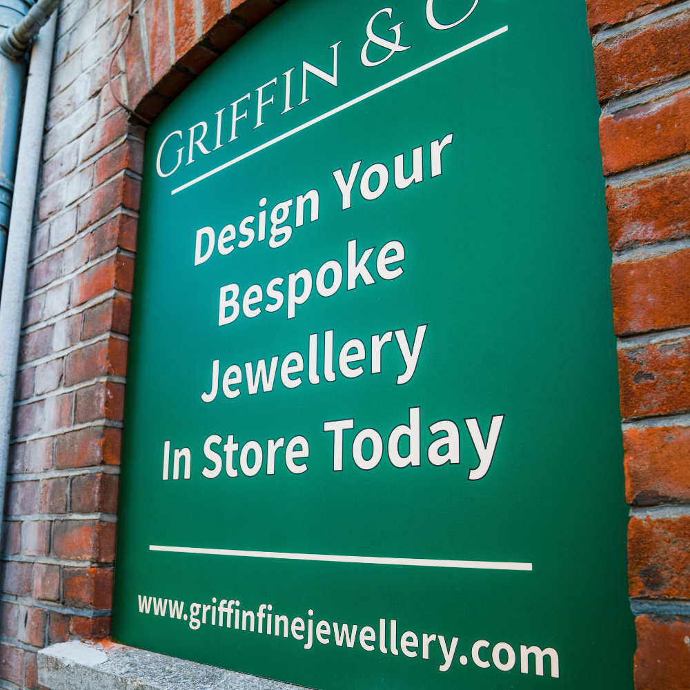 A close-up of a green sign with white text that reads, 'Design Your Bespoke Jewellery In Store Today,' followed by the website address for Griffin Fine Jewellery.