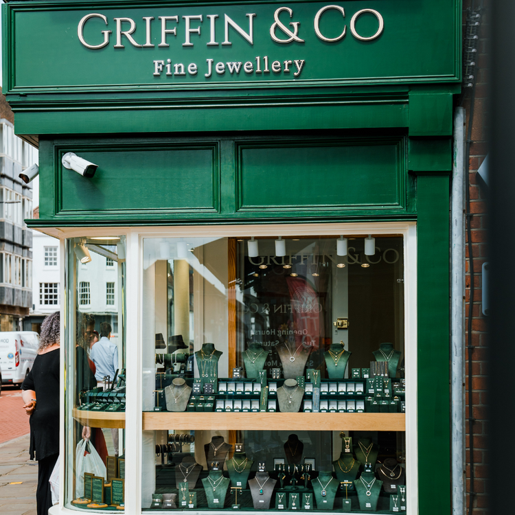A street view of the main display window of Griffin & Co. The large window is filled with several tiers of necklaces on display busts. A person stands on the pavement to the left of the window.