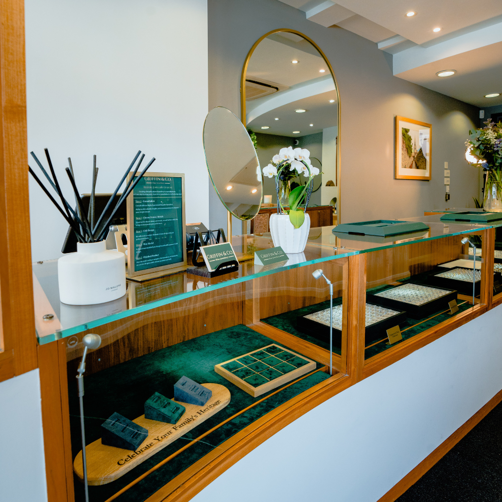 A view of the main display counter at Griffin & Co. Jewellery is displayed under the glass, while on top sits a decorative white orchid, a diffuser, and company literature. A large mirror and framed art hang on the wall behind.