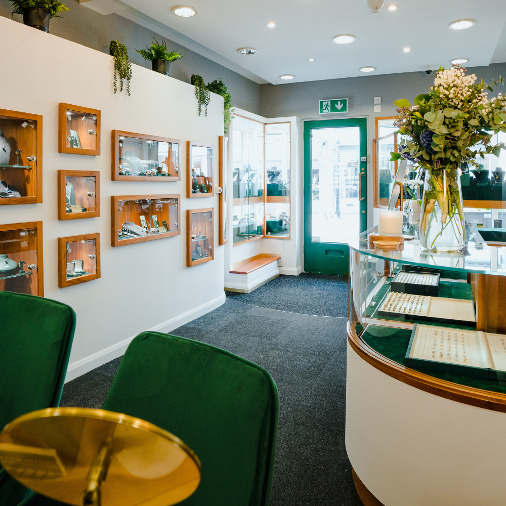 The bright and welcoming interior of the Griffin & Co showroom. The view shows a curved glass counter, green velvet consultation chairs, and jewellery displayed in wood-framed cases on the wall, with the entrance door in the distance.