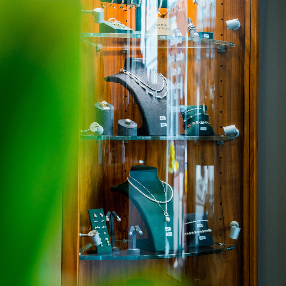 A view into a brightly lit display case containing necklaces, bracelets, and rings on two glass shelves. The pieces, including pearl necklaces, are arranged on black and grey display stands.