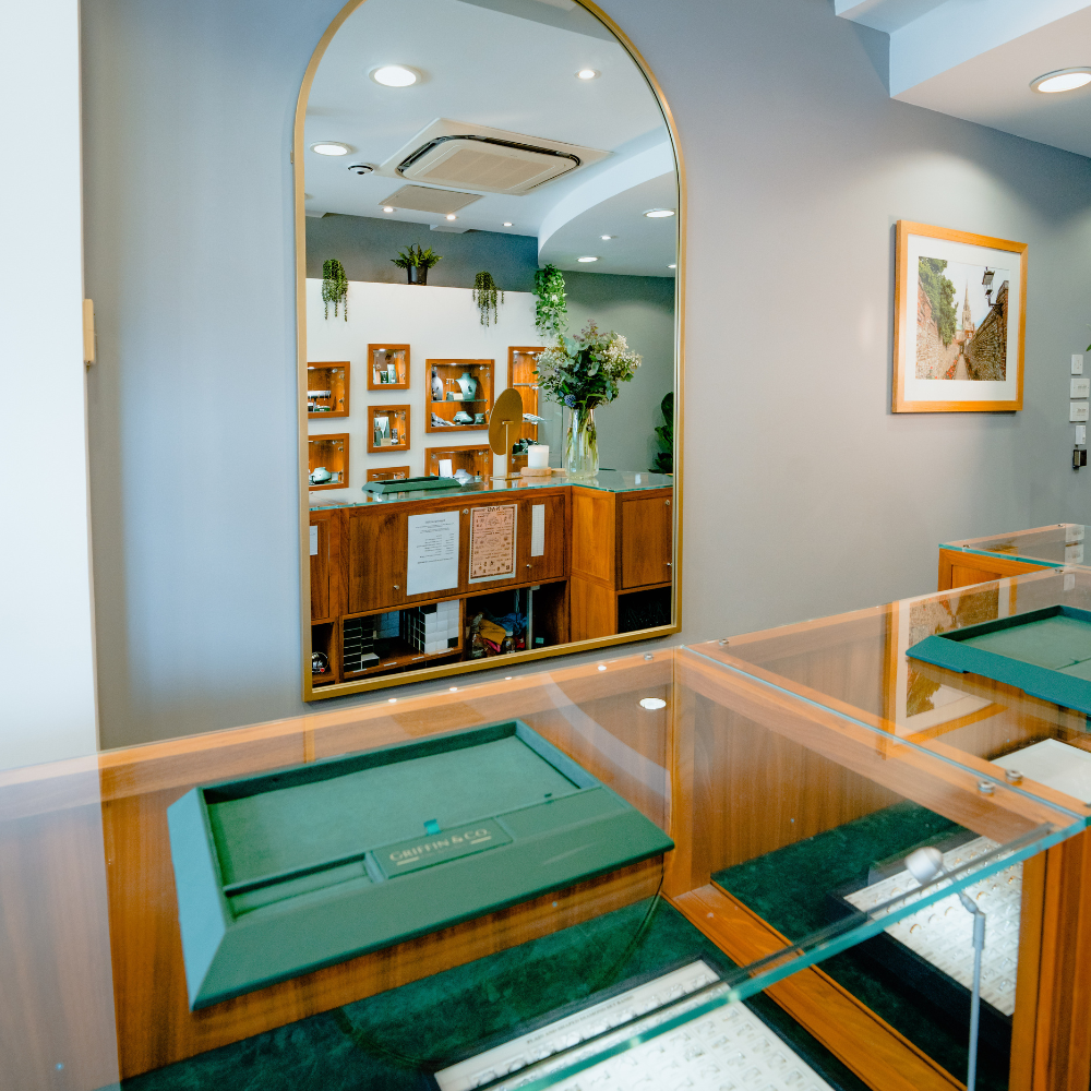 An interior shot of the showroom focusing on a large, arched mirror. The mirror reflects a clear image of the shop's counter, wall displays, and a large floral arrangement.