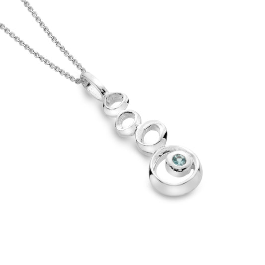 A product-only photo of the Silver Origins sterling silver pendant, featuring four stacked organic pebble shapes and a round blue topaz, on a white background. 1800