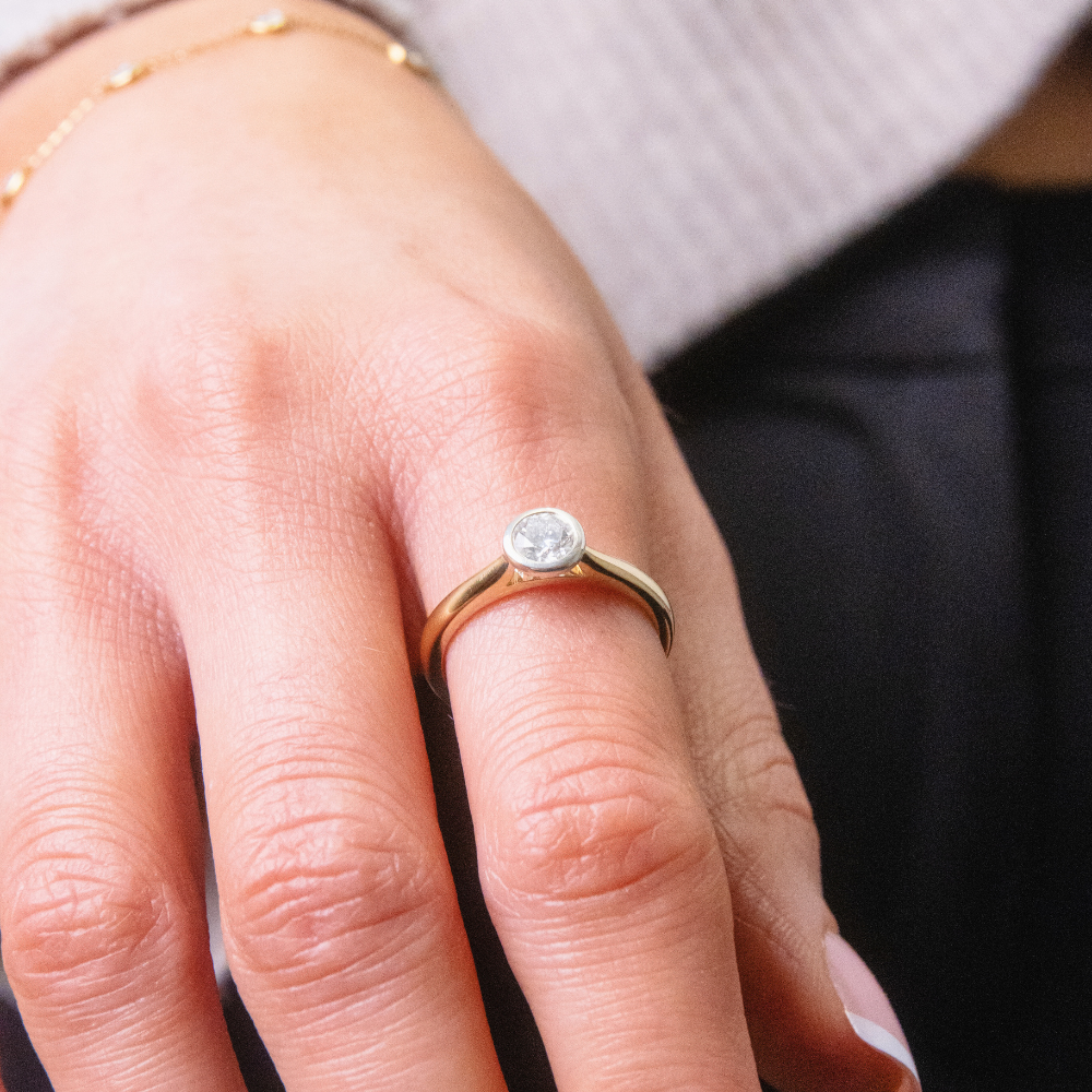 A woman's hand displays a modern and elegant lab-grown diamond solitaire ring with a smooth bezel setting in yellow gold, a bespoke ethical ring design from Griffin & Co Fine Jewellery.