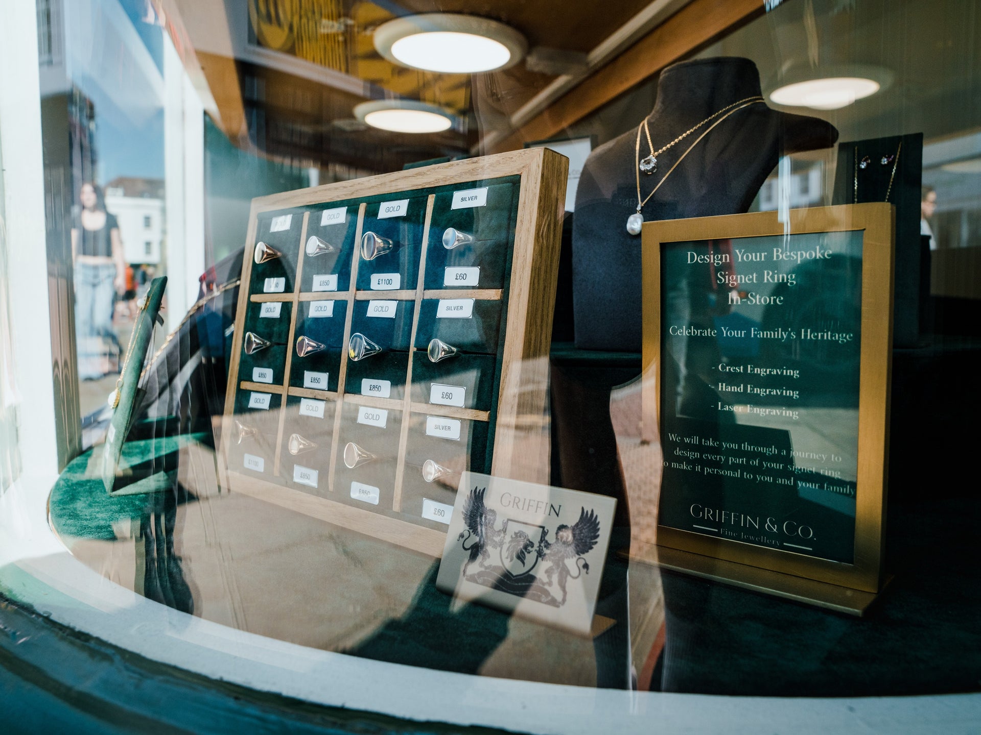 A view of a Griffin & Co. shop window from the outside. The display contains a variety of jewellery items, including necklaces on mannequins and a central stand of signet rings. A large sign with the text "Design Your Bespoke Signet Ring In-Store" is prominent, listing engraving services like "Crest Engraving" and "Hand Engraving."