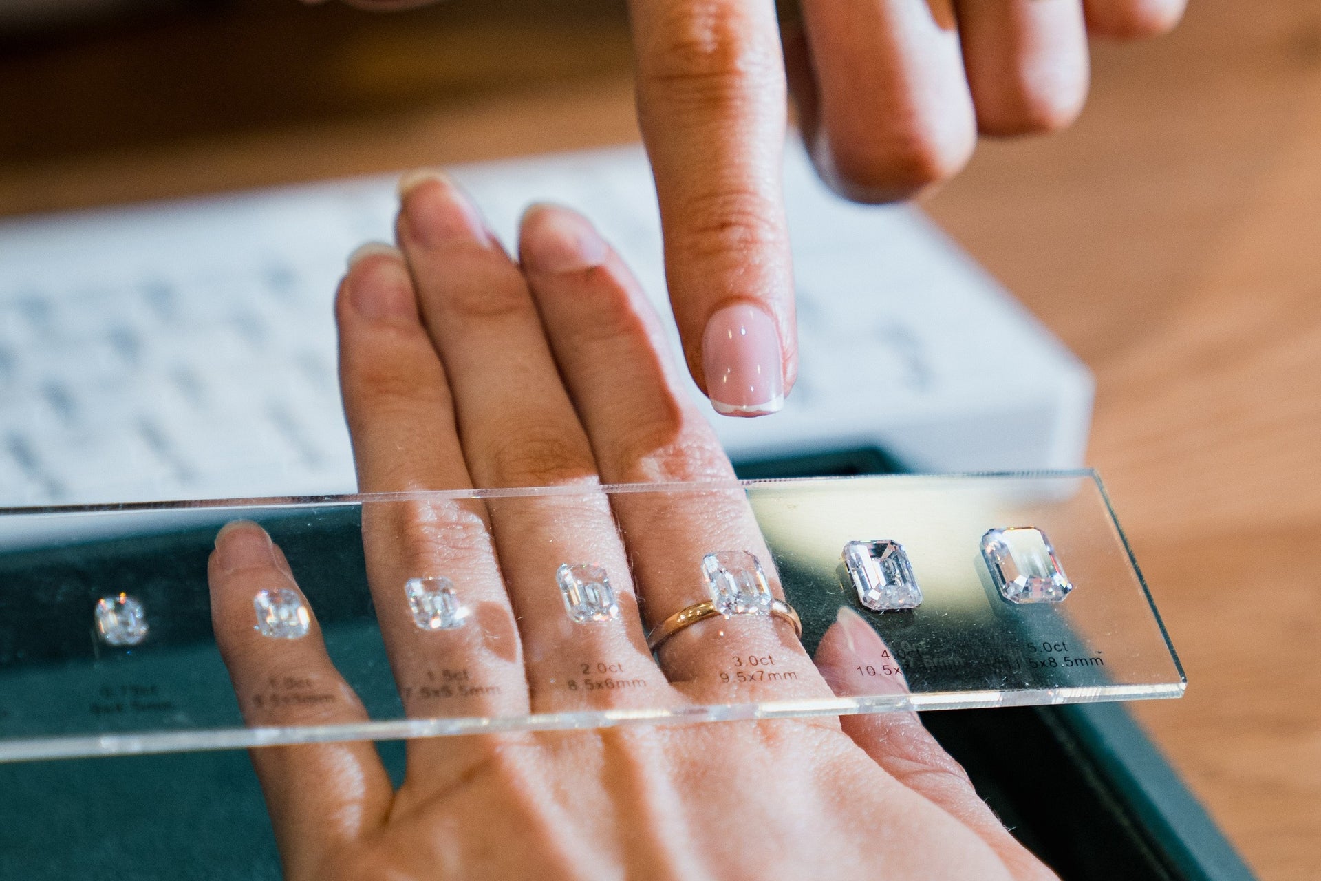 A close-up of a consultant pointing to a specific diamond size on a clear guide held over a customer's hand. This interactive tool helps demonstrate how different carat weights and shapes of lab-grown diamonds will look on the finger.