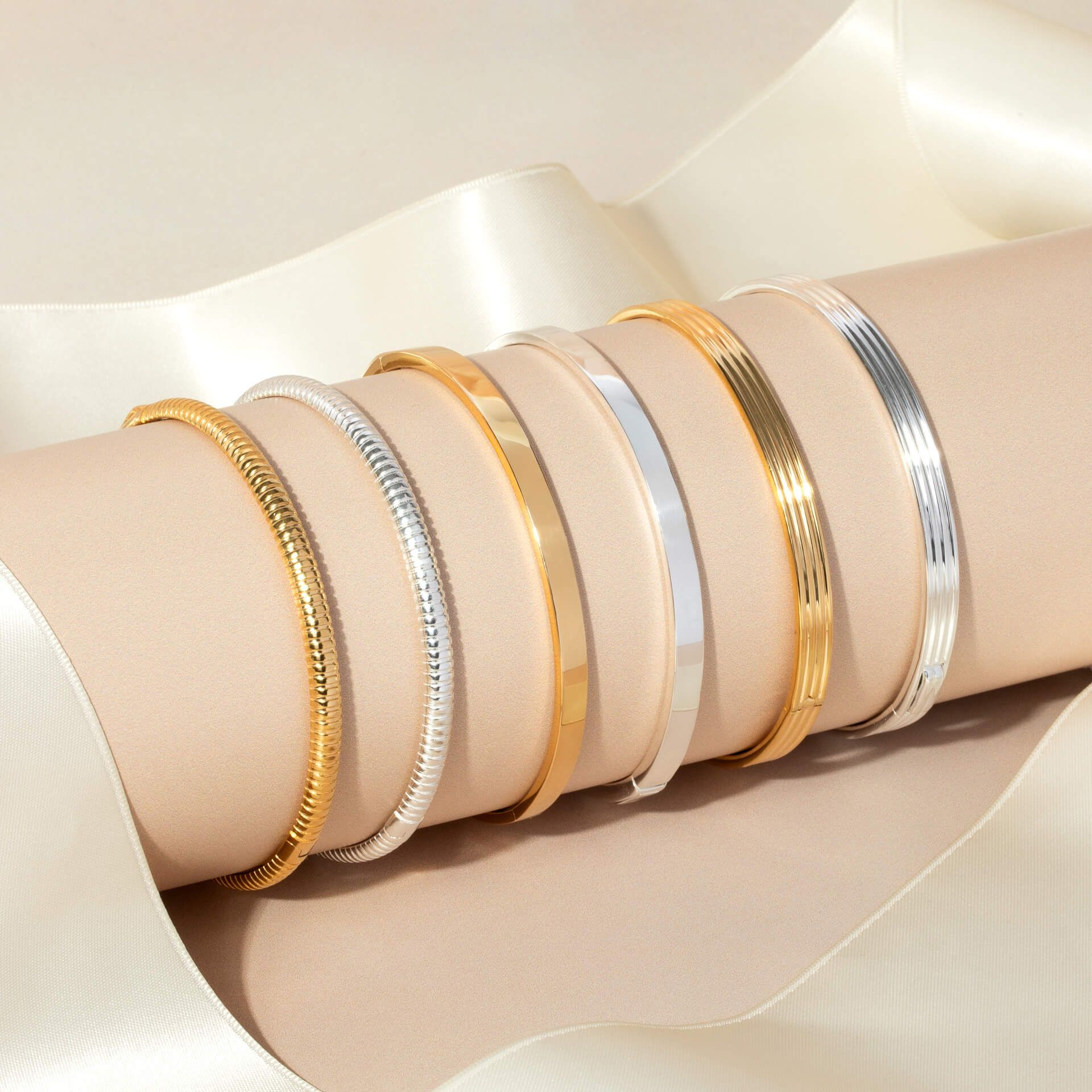 A collection of six stacking bangles in silver and gold, including two textured coil bangles, displayed on a beige cylinder with a cream ribbon background.