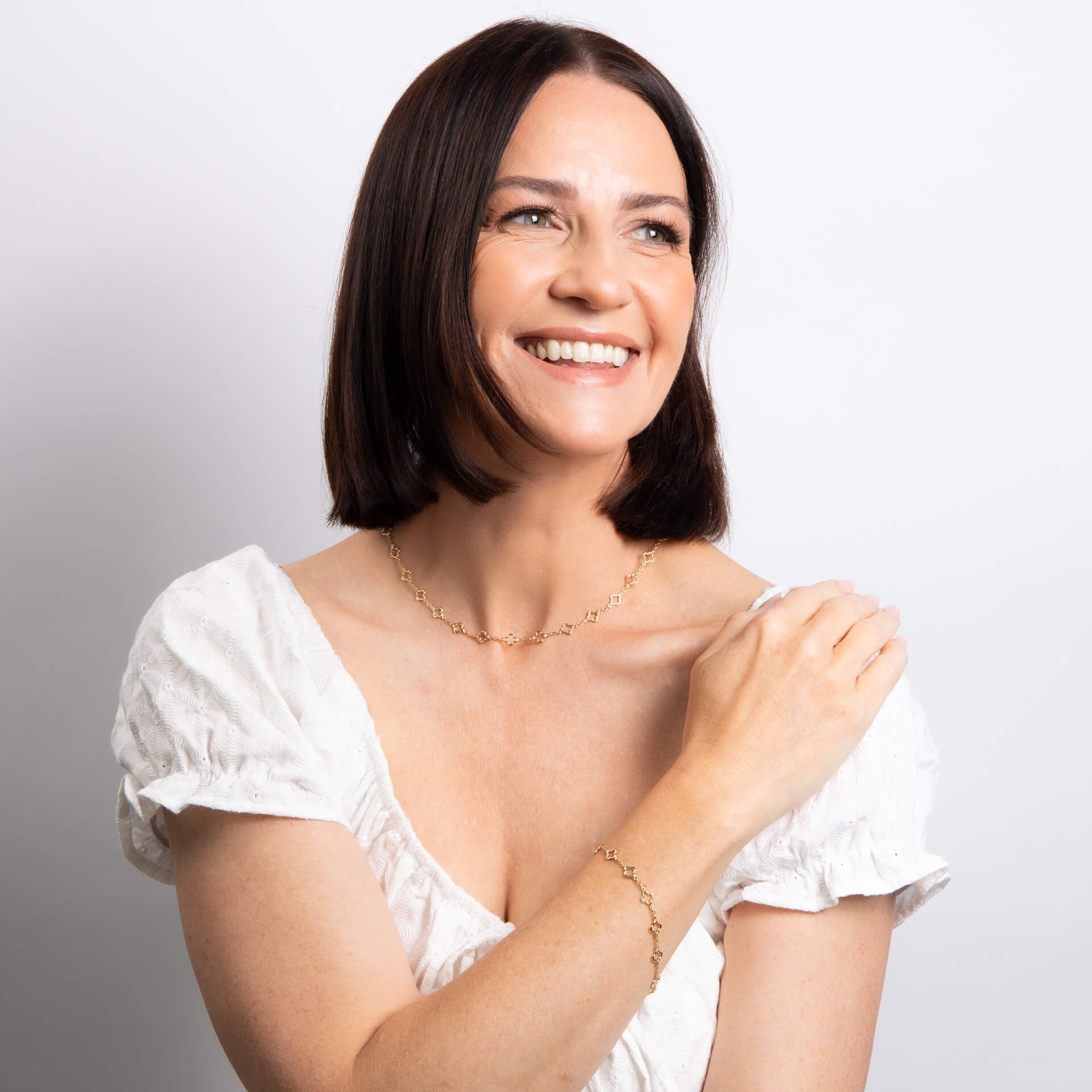 A smiling woman with a bob hairstyle wearing the gold quatrefoil station necklace and matching bracelet.