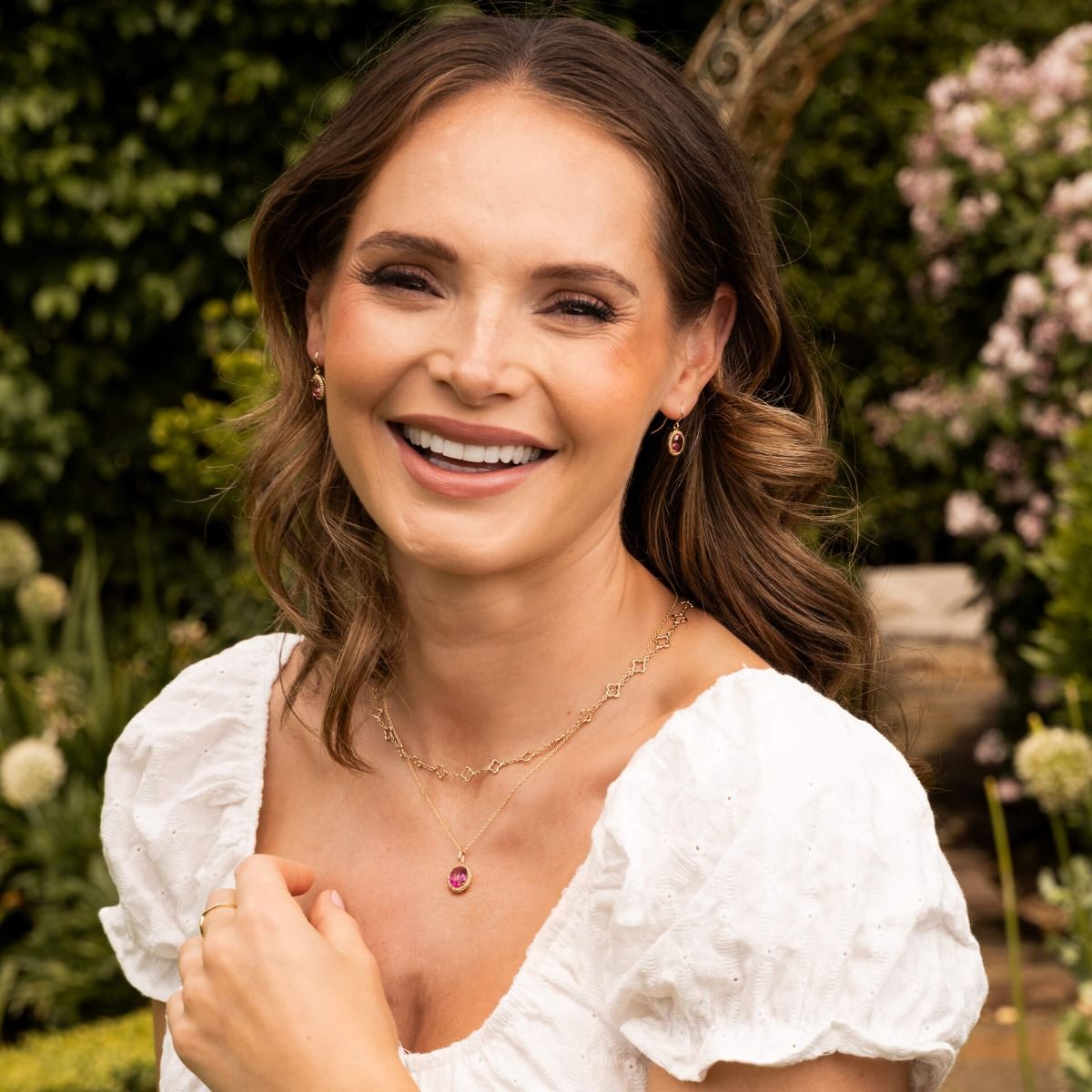 A smiling woman wearing the quatrefoil station necklace and bracelet layered with other gold and gemstone jewellery.