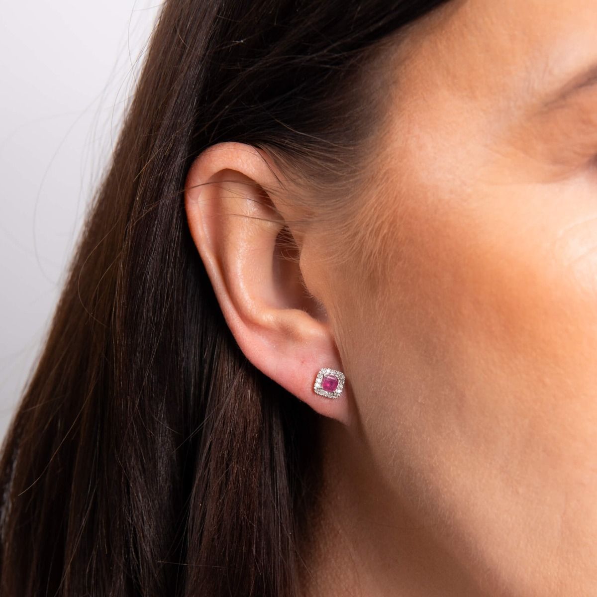 A close-up of a woman's ear wearing a ruby and diamond halo stud earring in white gold.
