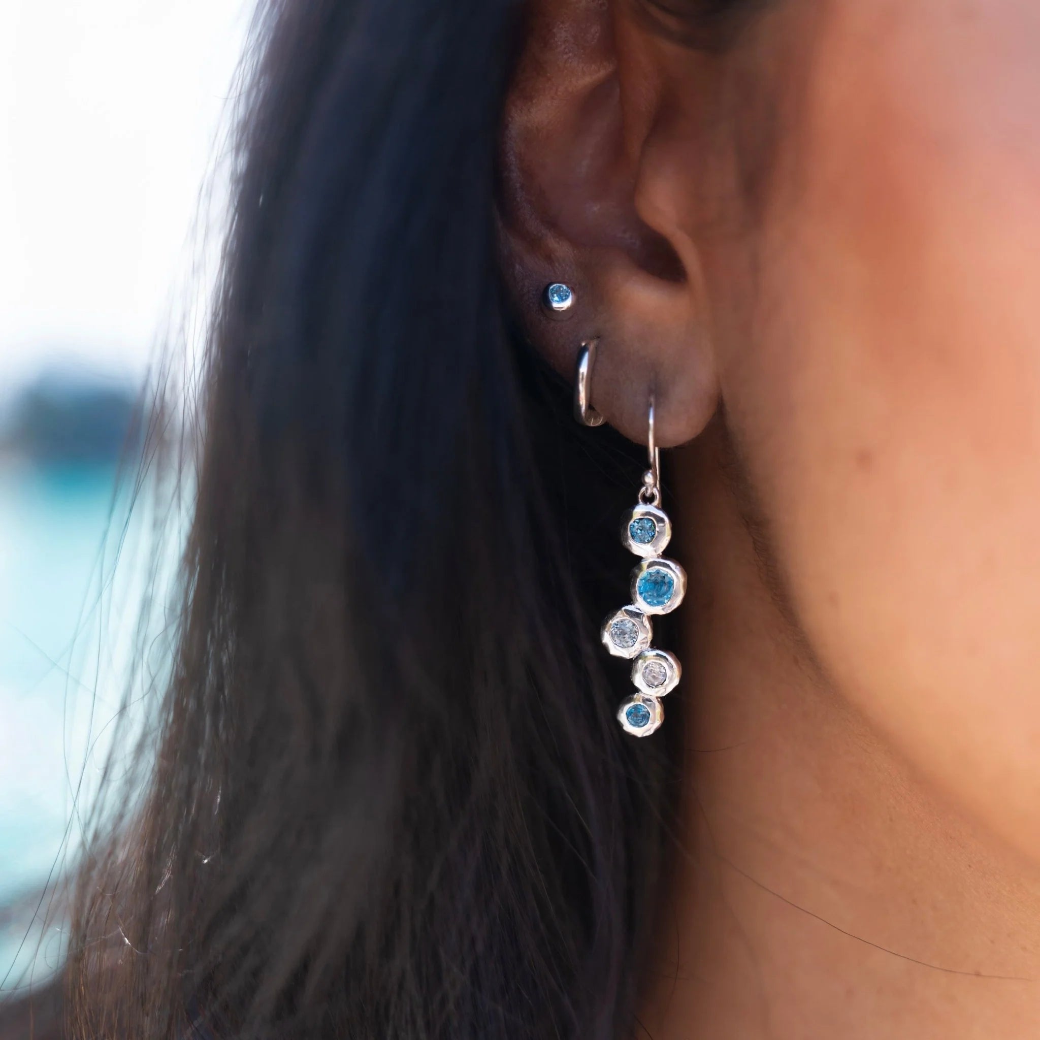 A close-up of a woman's ear and side of her face, with long dark hair. She is wearing one of the sterling silver topaz stream drop earrings, which hangs gracefully below her lobe. Other small silver earrings are also visible in her ear.