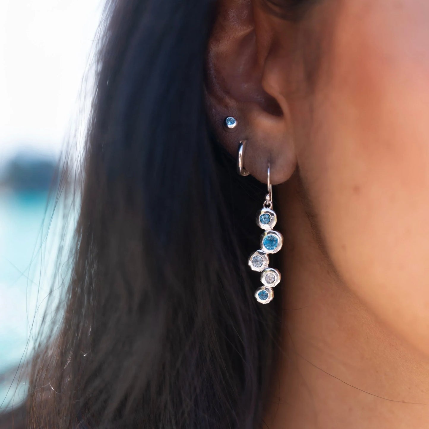 A close-up of a woman's ear and side of her face, with long dark hair. She is wearing one of the sterling silver topaz stream drop earrings, which hangs gracefully below her lobe. Other small silver earrings are also visible in her ear.