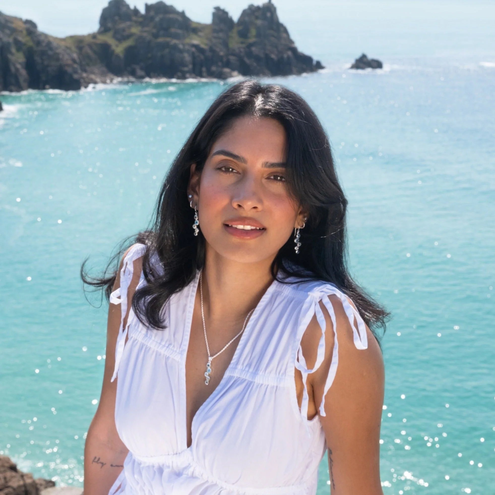 A woman with long dark hair, wearing a white v-neck top with tie-up sleeves, smiling at the camera. She is standing outdoors with a bright blue sea and rocky coastline in the background, wearing the sterling silver topaz stream drop earrings and a matching pendant.